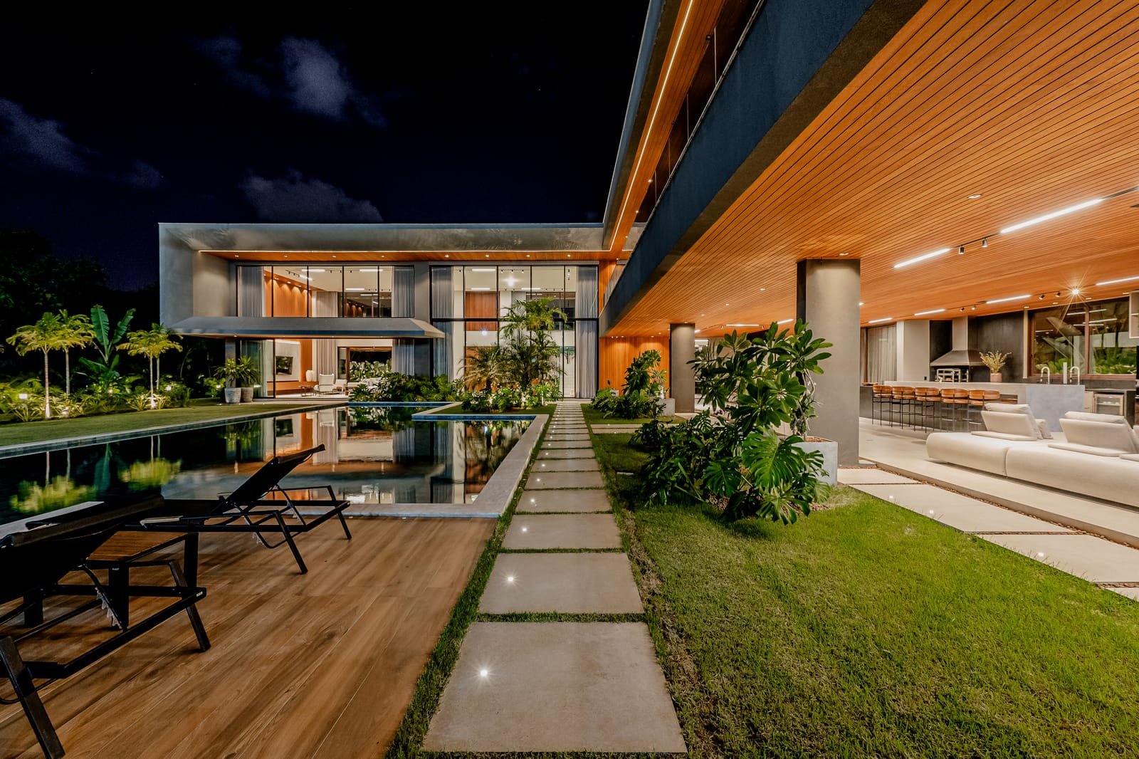 Sala de estar aberta para o jardim por uma faixa contínua de vidro, com forro de madeira e vegetação rente à fachada.