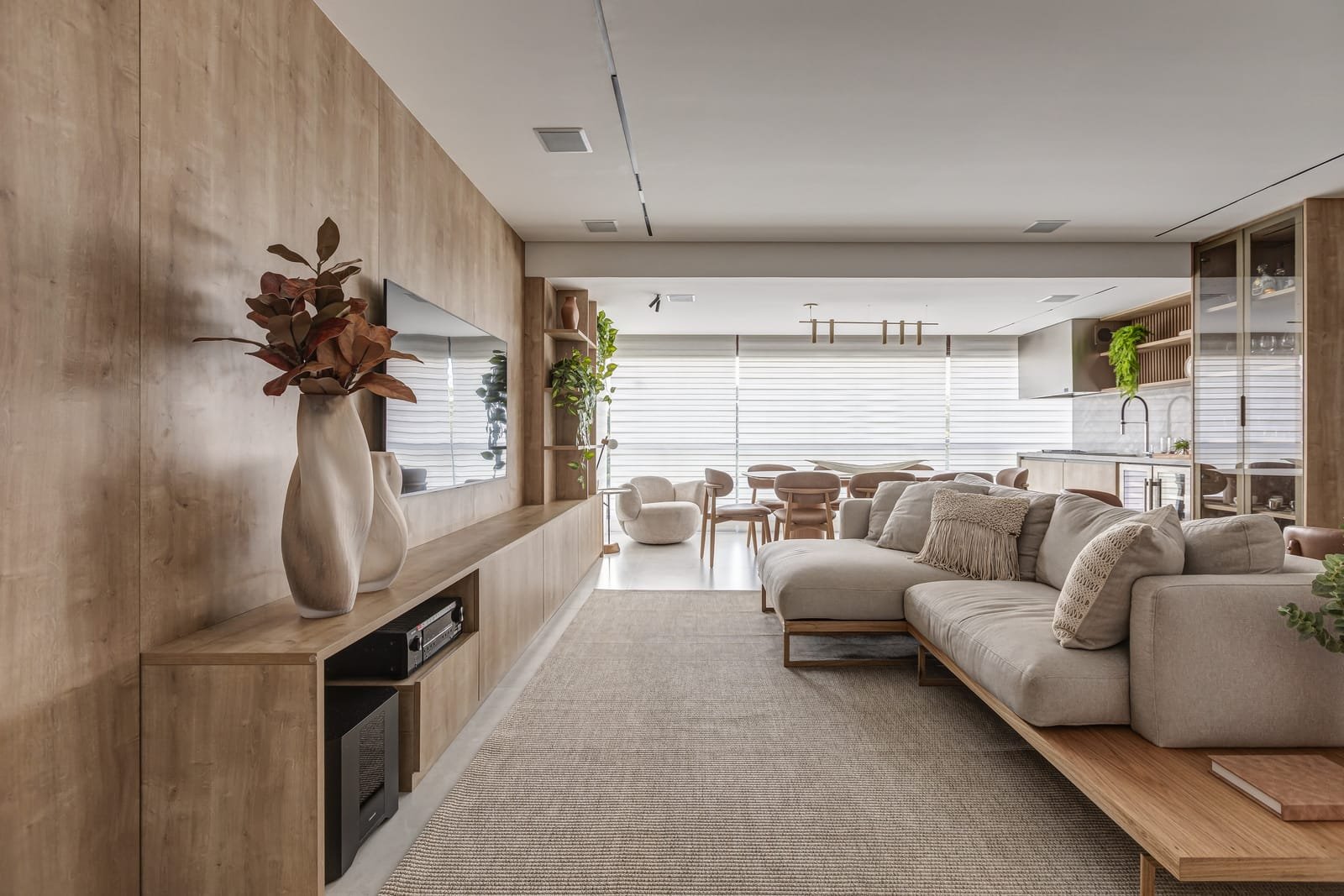 Living e sala de jantar de apartamento em enfiada, com marcenaria de madeira clara, plantas pendentes e persianas filtrando a luz.