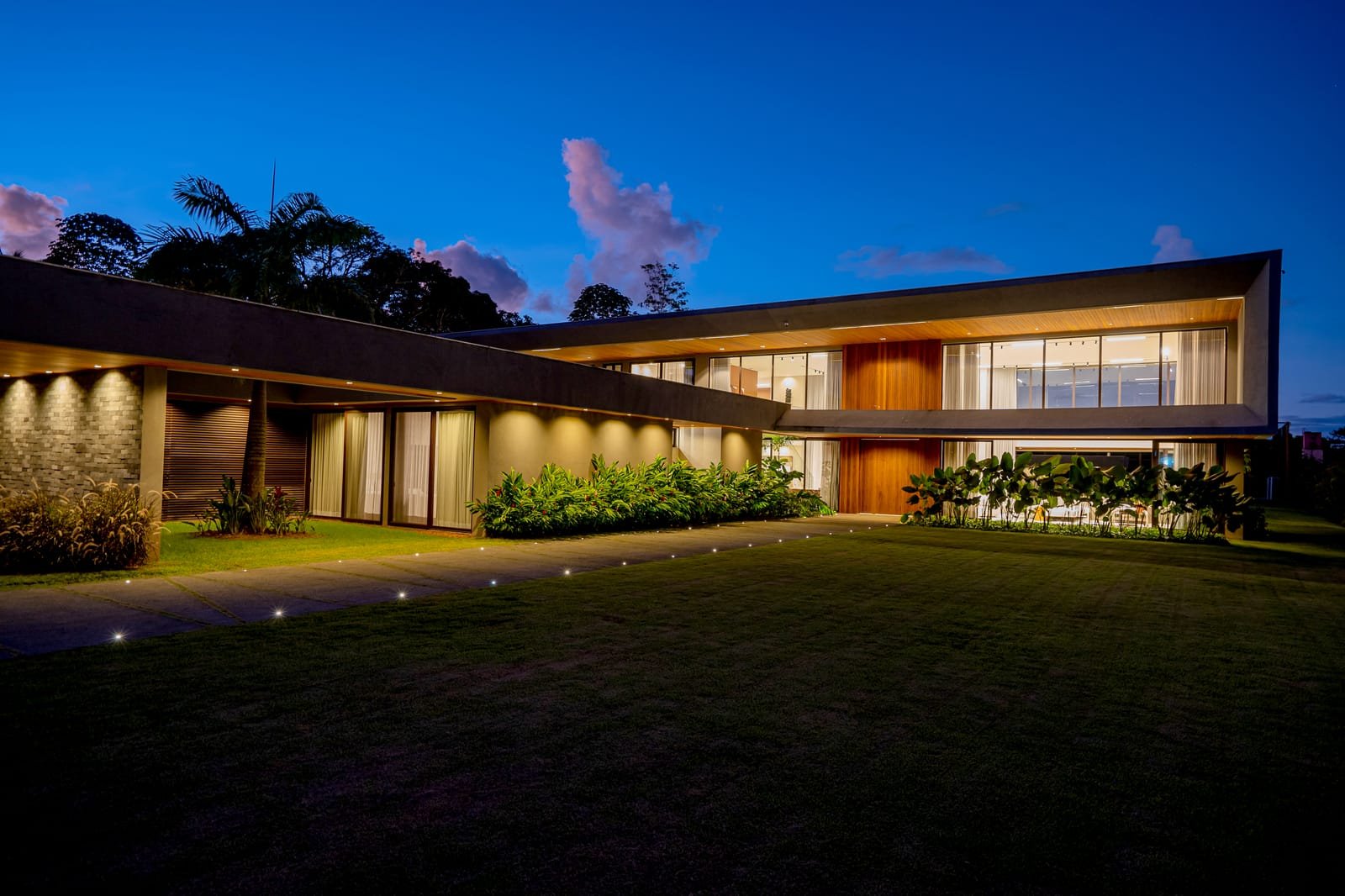 Casa de alto padrão na hora azul: volume horizontal de concreto deitado sobre o gramado, interiores acesos atrás dos panos de vidro e balizadores marcando o caminho.