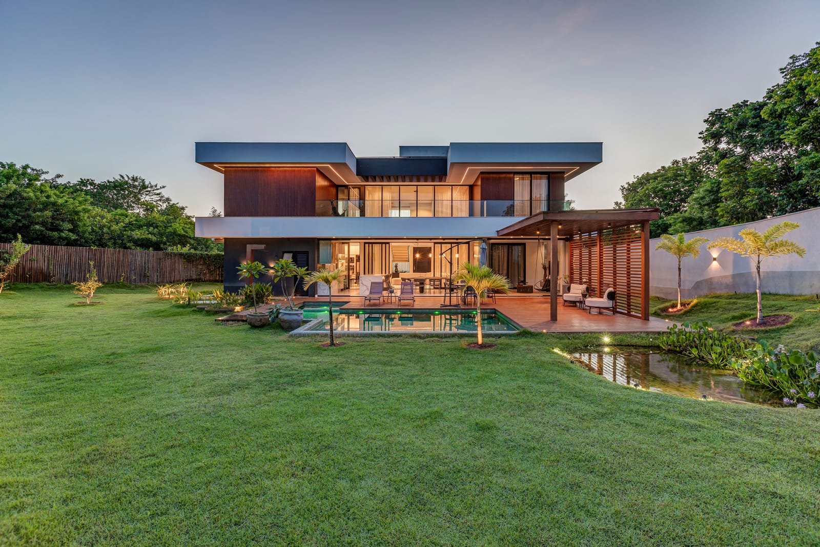 Casa térrea vista do gramado ao entardecer, com piscina, deck de madeira e brises verticais na varanda.
